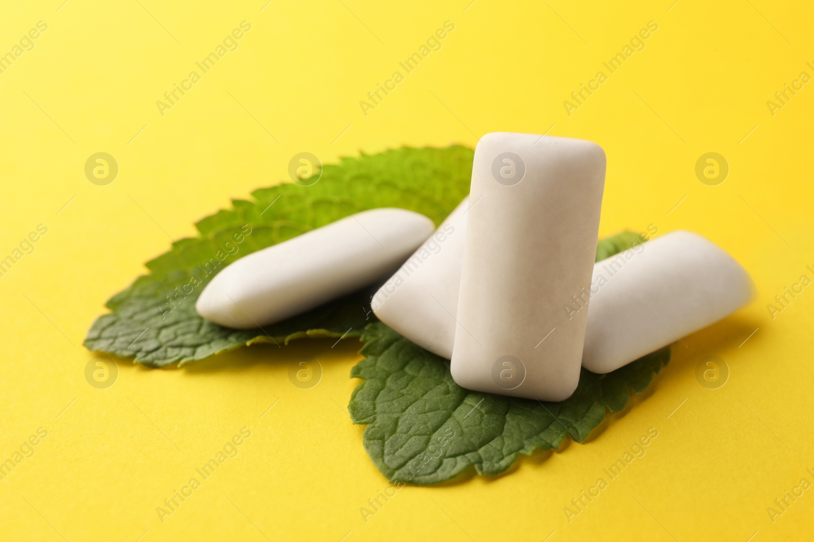Photo of Tasty white chewing gums and mint leaves on yellow background, closeup