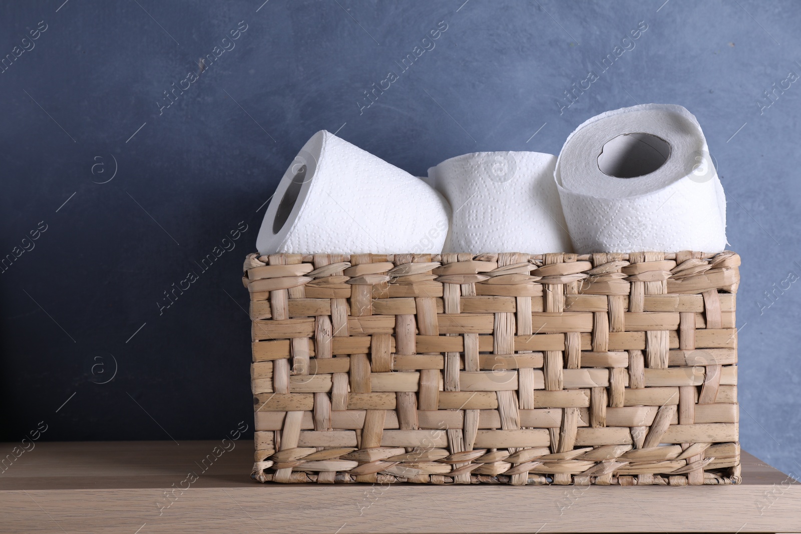 Photo of Toilet paper rolls in wicker basket on wooden table near blue wall