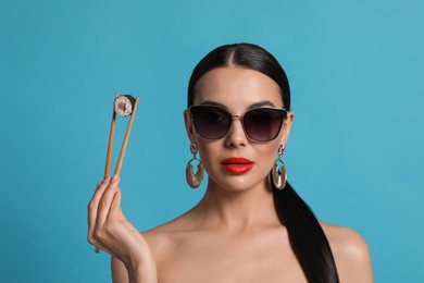 Photo of Attractive woman in fashionable sunglasses holding chopsticks with sushi against light blue background