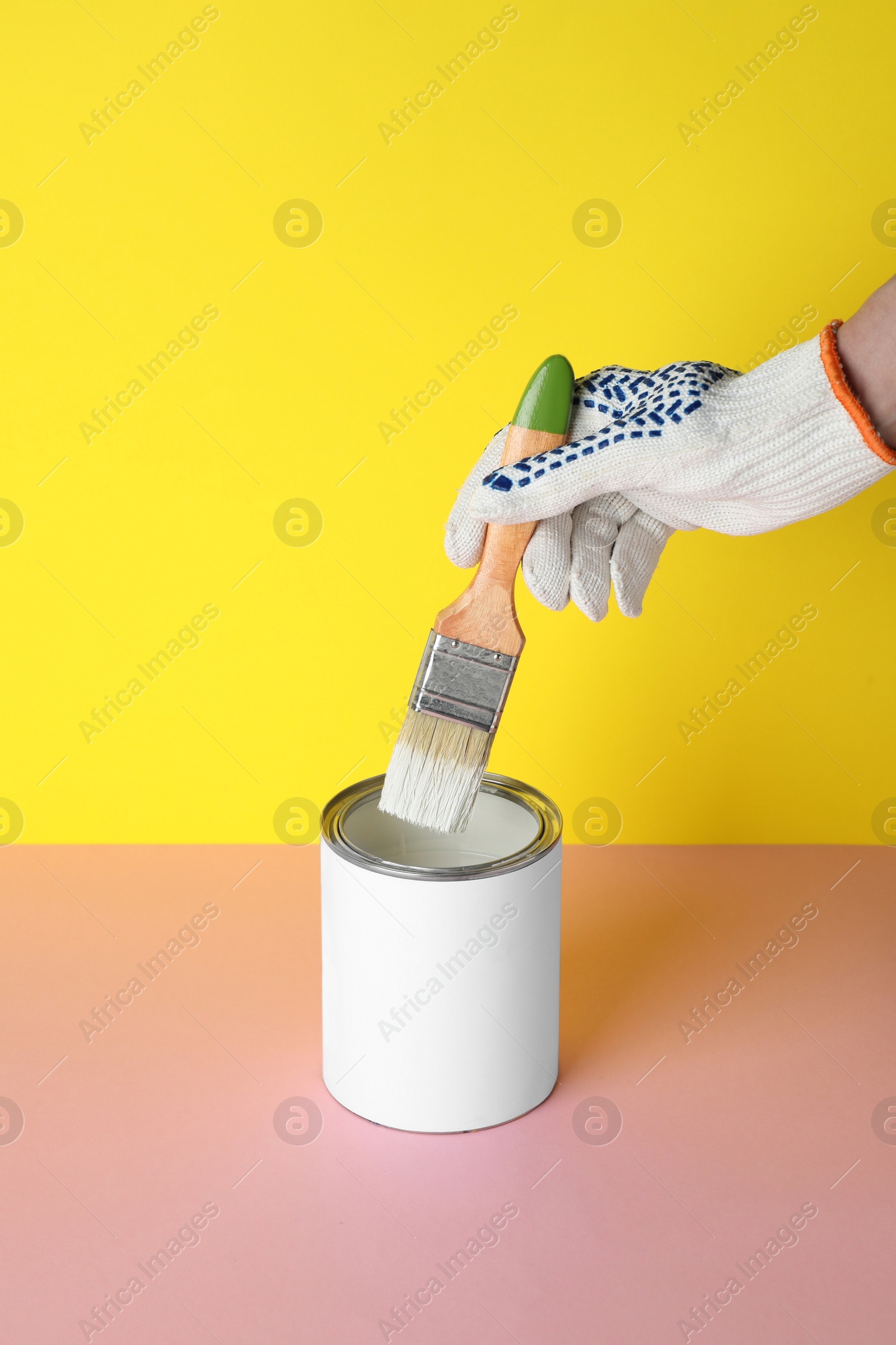 Photo of Person dipping brush into can of white paint on pink table against yellow background, closeup. Mockup for design