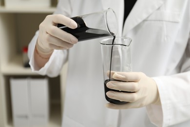 Photo of Laboratory worker pouring black crude oil into flask indoors, closeup