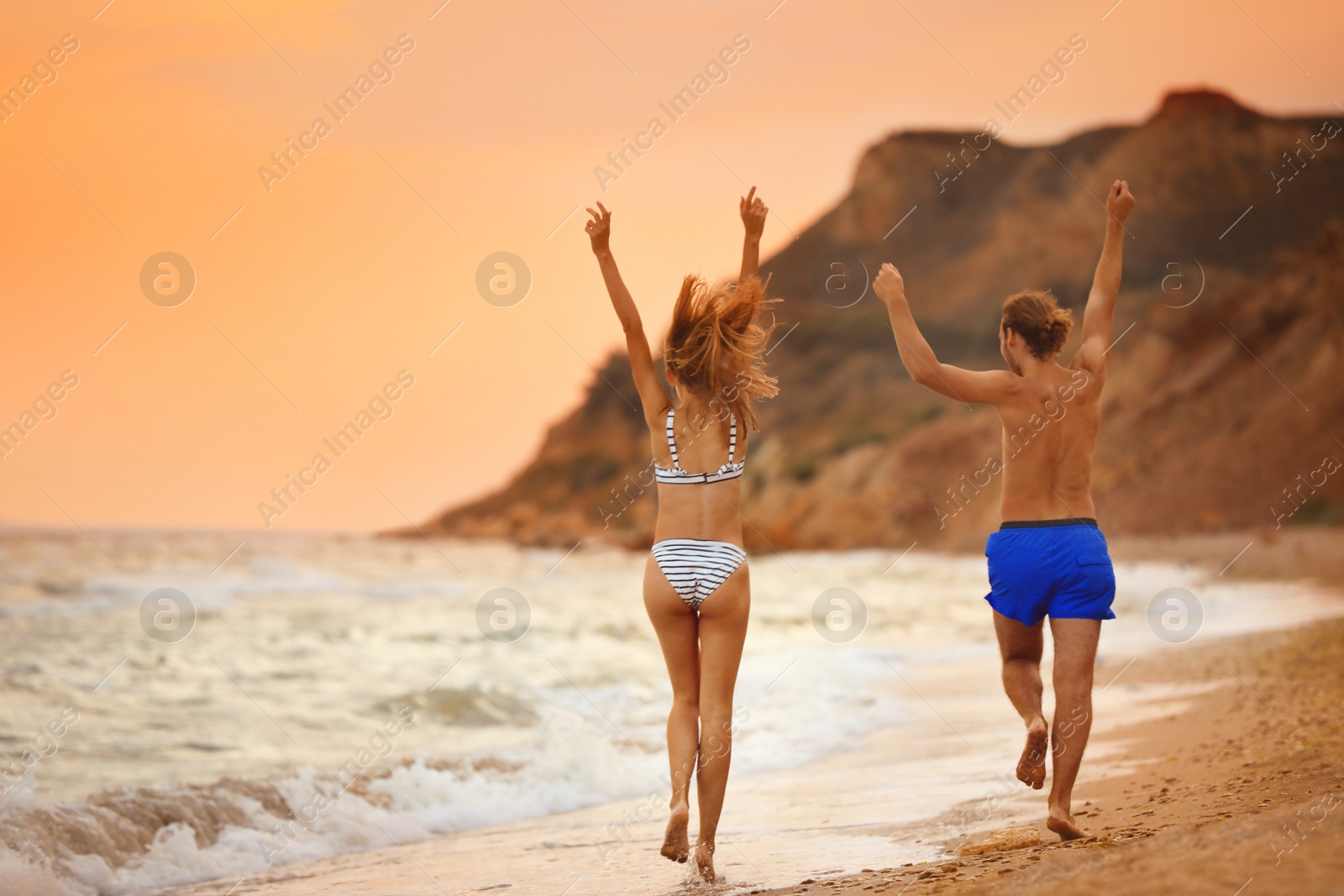 Photo of Young woman in bikini and her boyfriend walking on beach at sunset. Lovely couple