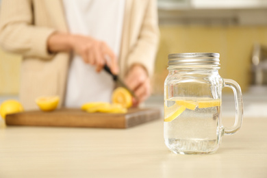 Photo of Woman cutting fruits in kitchen, focus on mason jar with lemon water