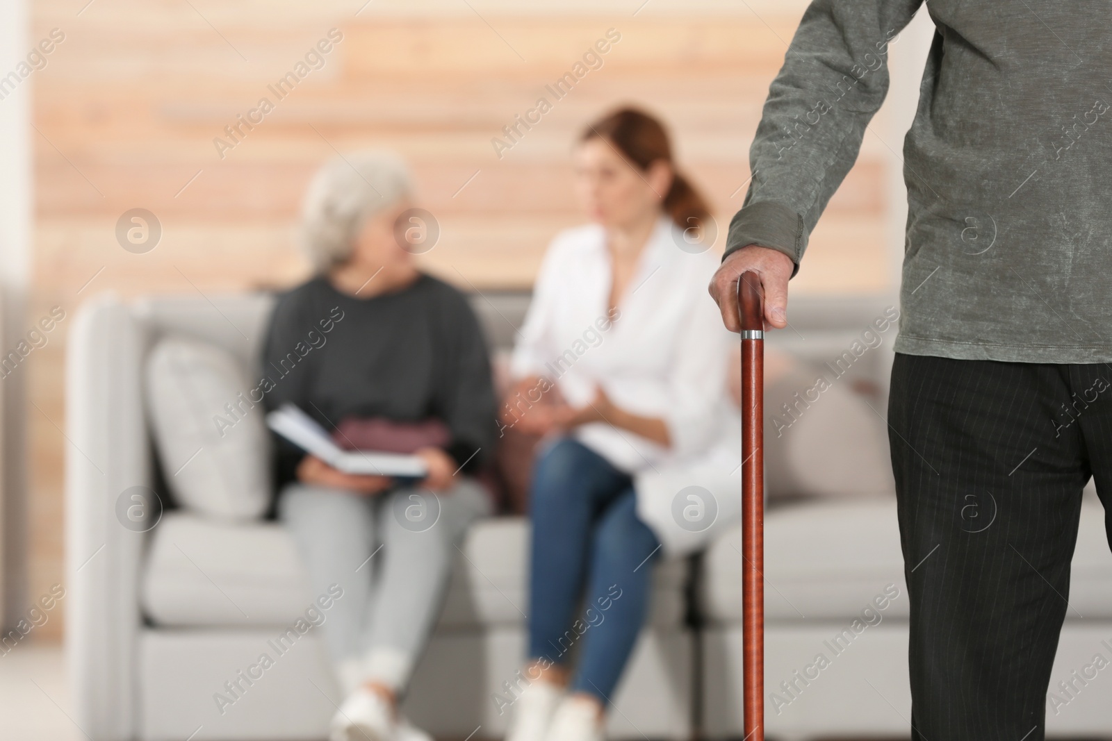 Photo of Elderly man holding walking cane and blurred caregiver with senior woman on background, focus on hand. Space for text