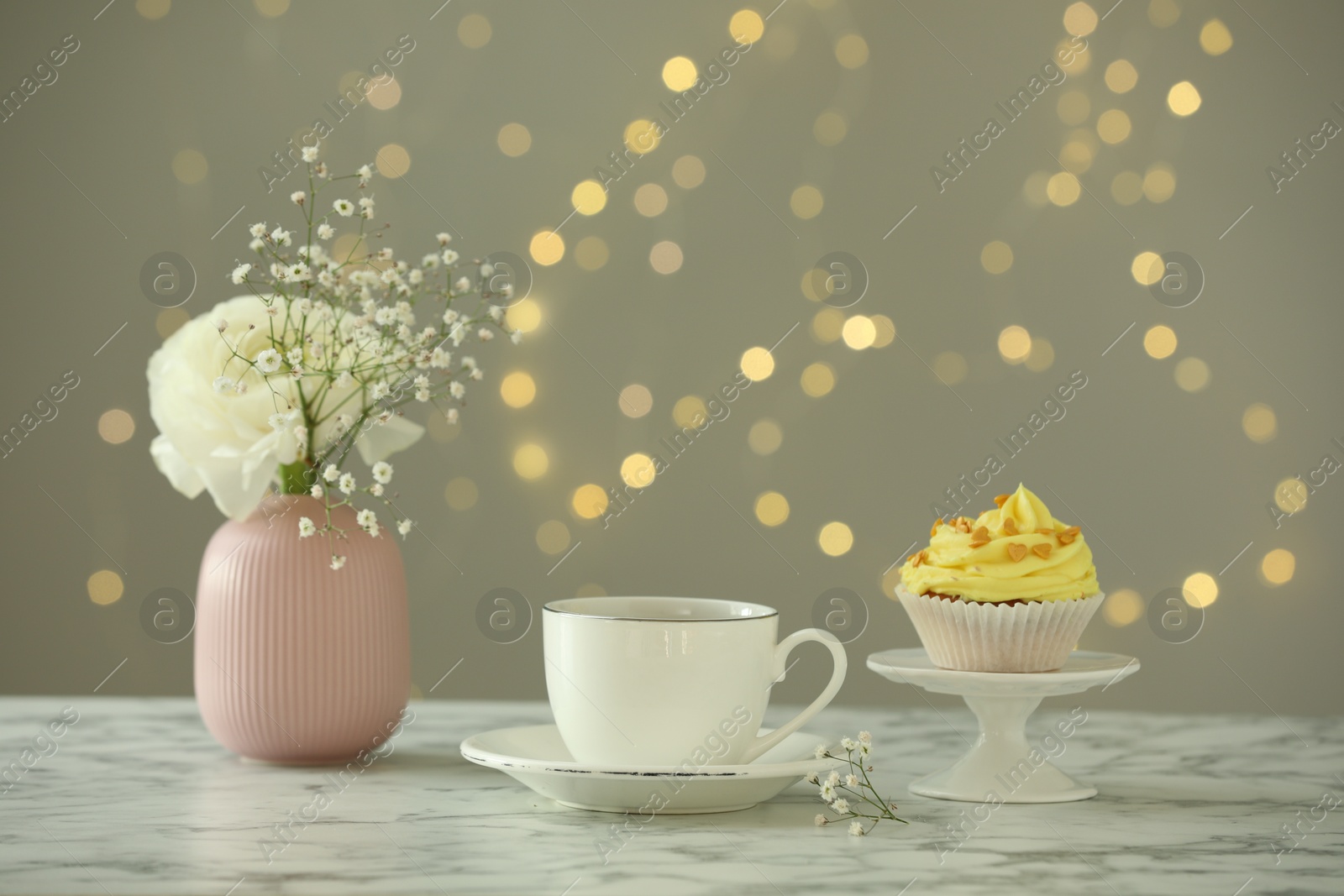 Photo of Delicious cupcake with yellow cream, tea and flowers on white marble table against blurred lights