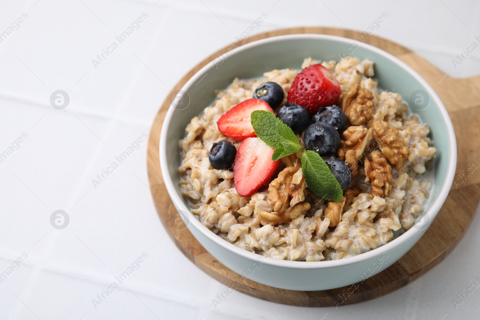 Photo of Tasty oatmeal with strawberries, blueberries and walnuts in bowl on white tiled table, closeup. Space for text