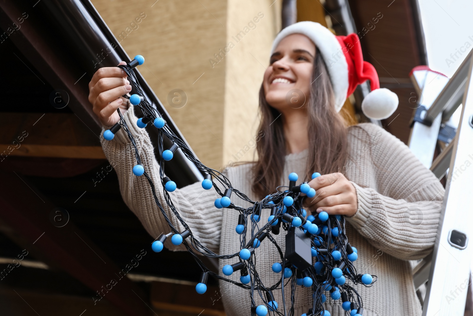 Photo of Happy woman in Santa hat decorating house with Christmas lights outdoors, selective focus