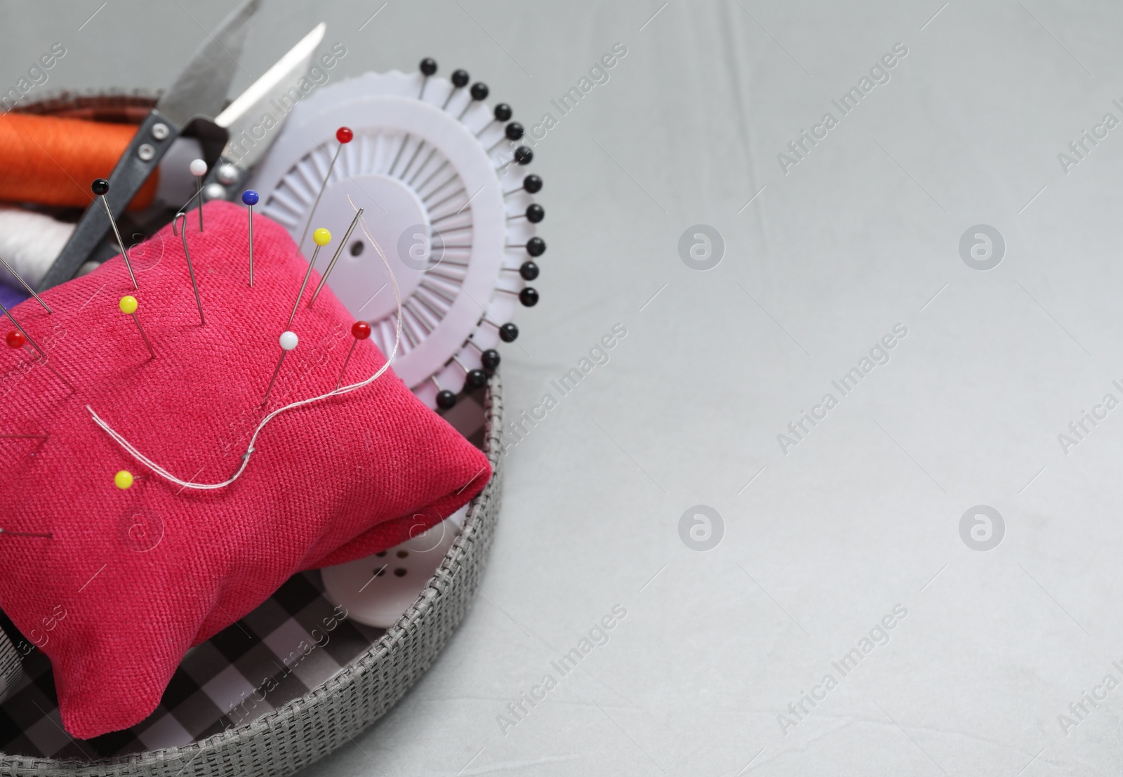 Photo of Red pincushion with pins and other sewing tools on grey table, closeup. Space for text