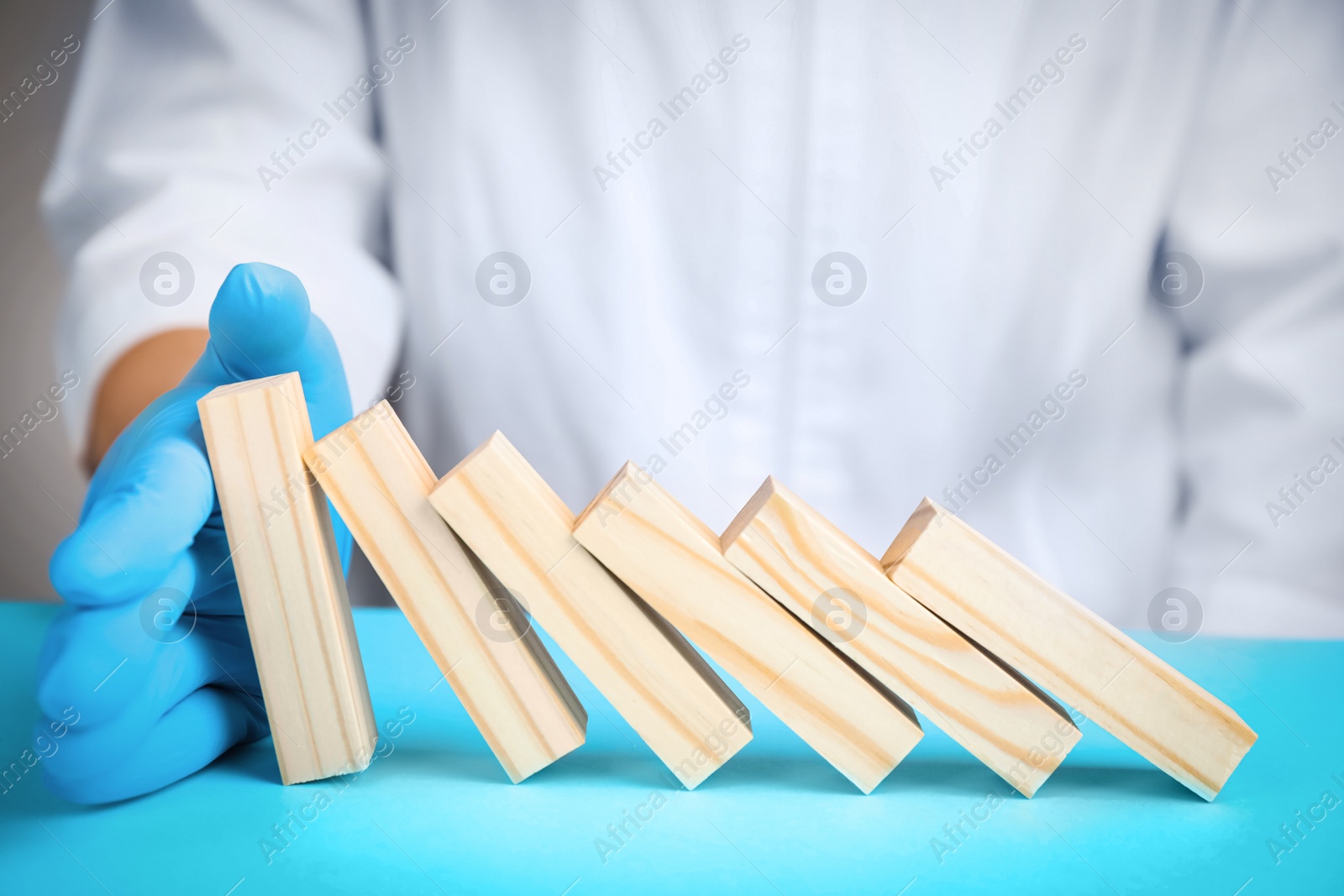 Photo of Doctor stopping domino effect at light blue table, closeup. Prevent spreading of coronavirus
