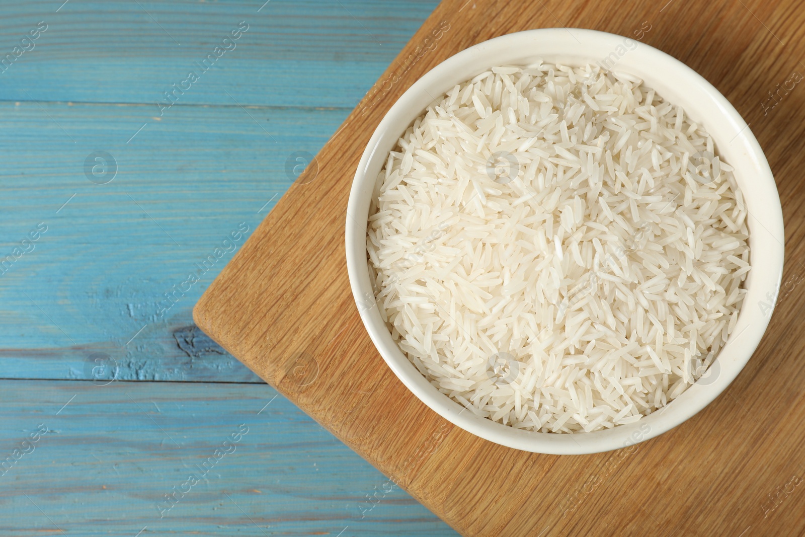 Photo of Raw basmati rice in bowl on light blue wooden table, top view. Space for text