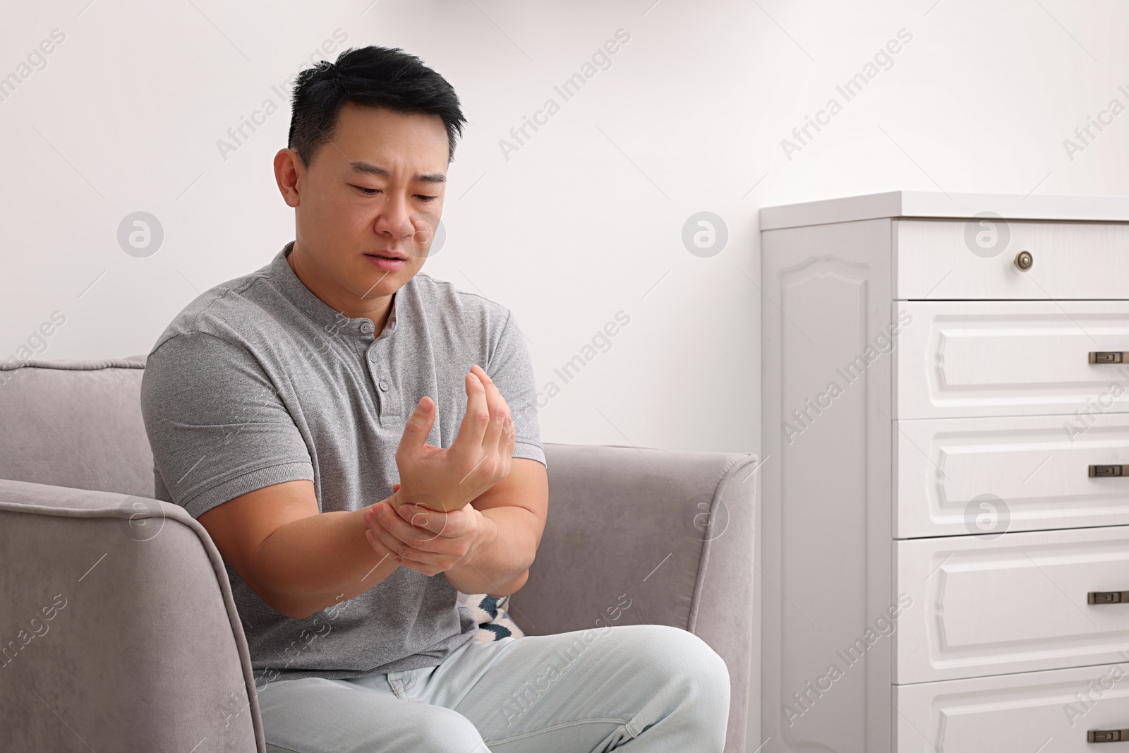 Photo of Asian man suffering from pain in his hand on armchair indoors