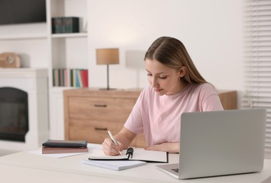 Photo of Online learning. Teenage girl writing in notepad near laptop at table