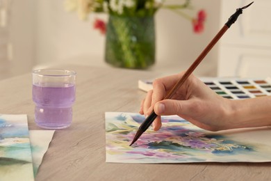 Photo of Woman painting flowers with watercolor at white wooden table indoors, closeup. Creative artwork