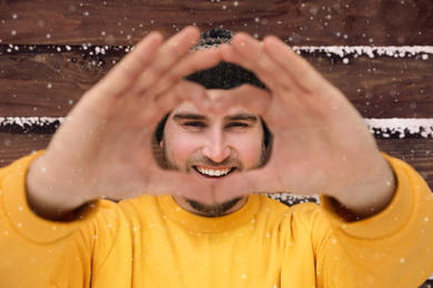 Photo of Young man making heart with his hands near wooden wall.  Winter vacation