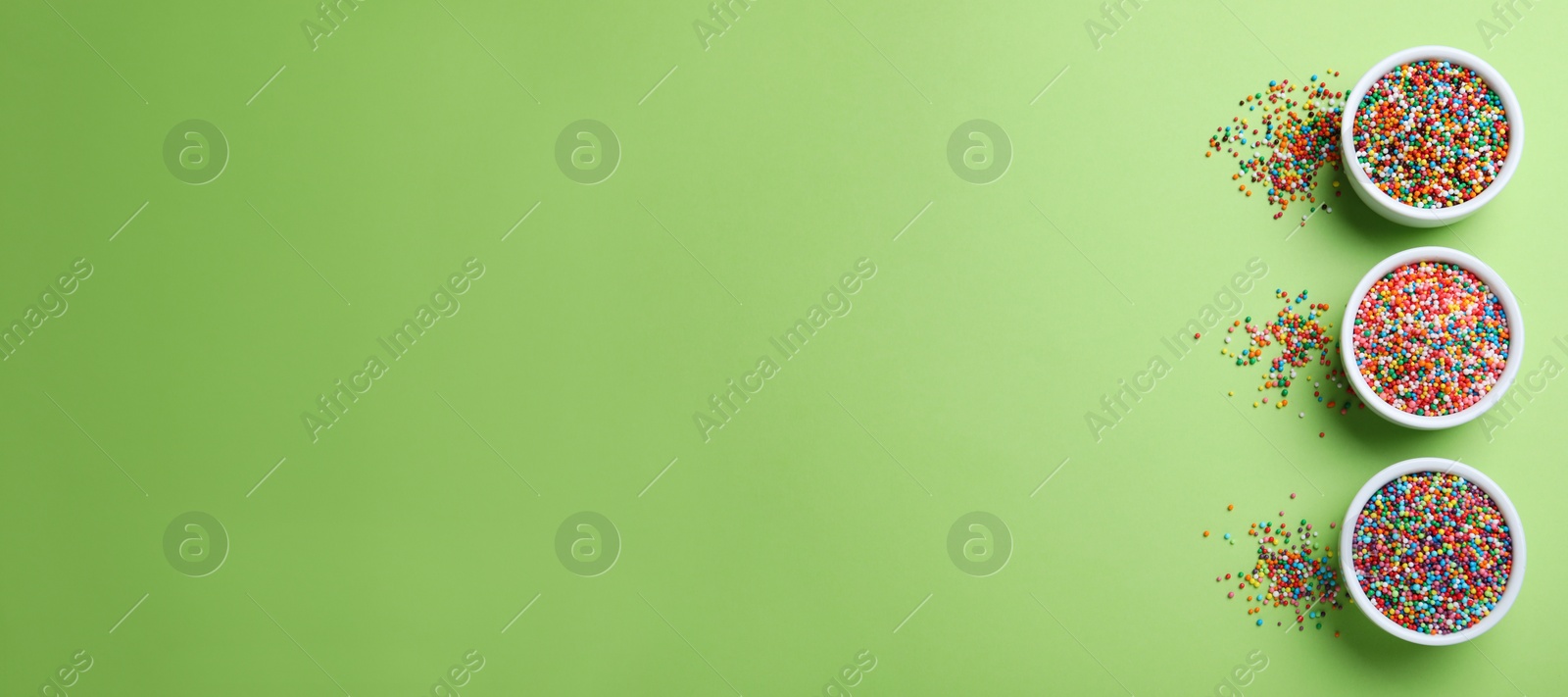 Photo of Colorful sprinkles and bowls on green background, flat lay with space for text. Confectionery decor