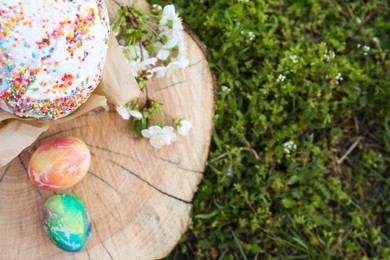 Photo of Easter kulich and painted eggs near blossoming tree twigs on wooden stump outdoors, top view. Space for text