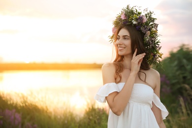 Photo of Young woman wearing wreath made of beautiful flowers outdoors at sunset