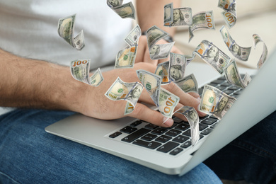 Man with modern laptop and flying dollar banknotes indoors, closeup. People make money online