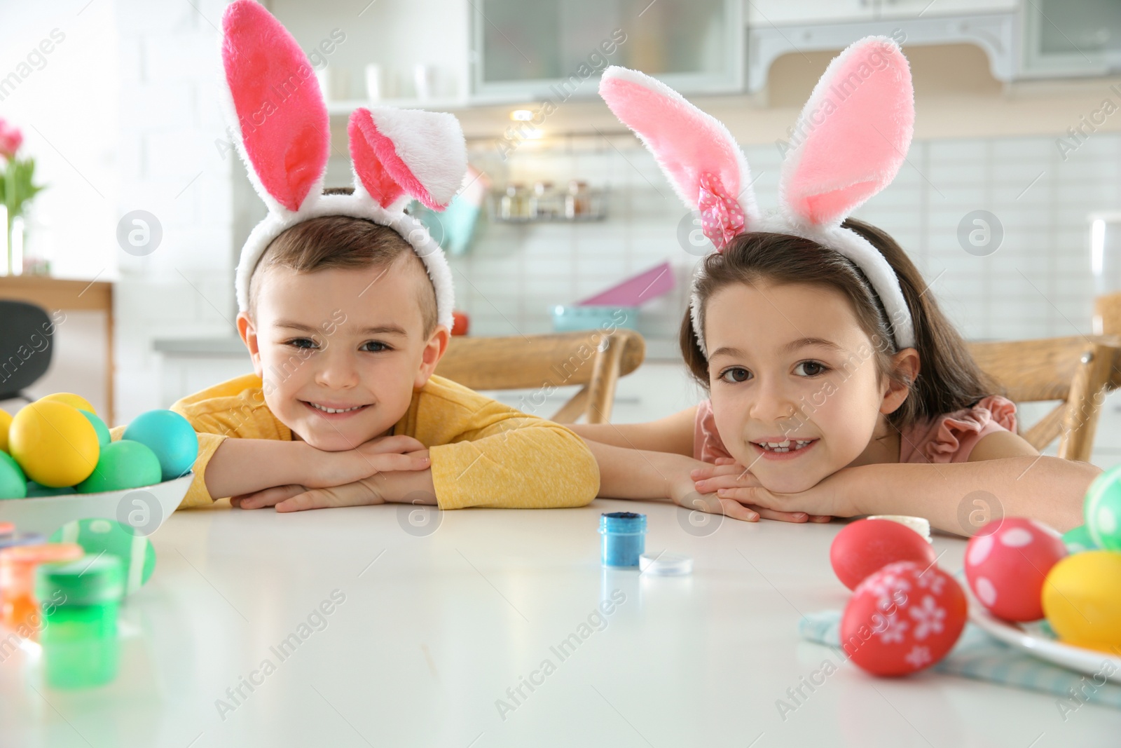 Photo of Cute children with bunny ears headbands and painted Easter eggs sitting at table in kitchen
