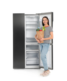 Young woman with bag of groceries near open empty refrigerator on white background