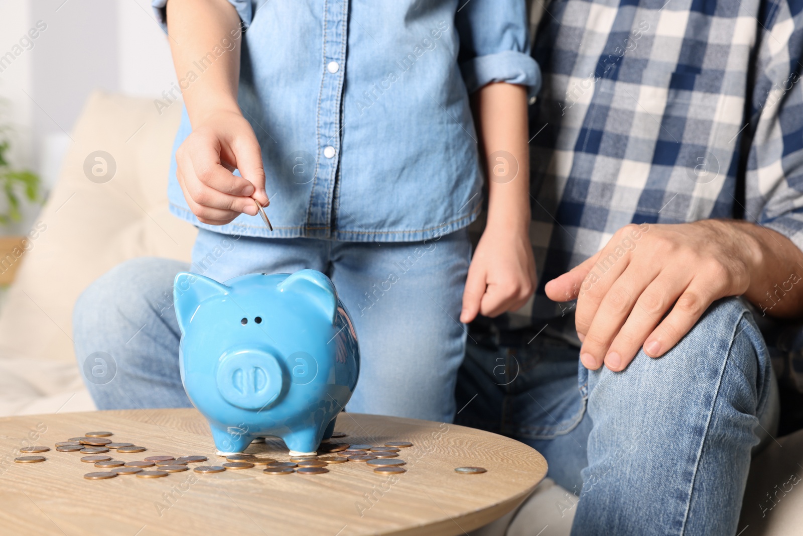 Photo of Little girl with her father putting coin into piggy bank at home, closeup