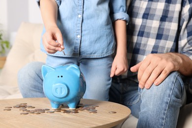 Little girl with her father putting coin into piggy bank at home, closeup