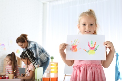 Photo of Cute little child showing sheet of paper with colorful hand prints indoors. Painting lesson