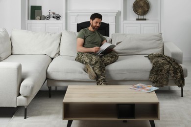 Photo of Happy soldier reading magazine on sofa in living room. Military service
