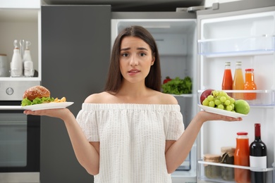 Photo of Doubtful woman choosing between fruits and burger with French fries near refrigerator in kitchen