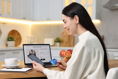 Beautiful young woman with credit card and gadgets shopping online at wooden table in kitchen