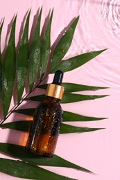 Bottle of cosmetic oil and tropical leaf in water on pink background, flat lay