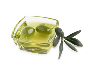 Photo of Glass bowl of oil, ripe olives and green leaves on white background