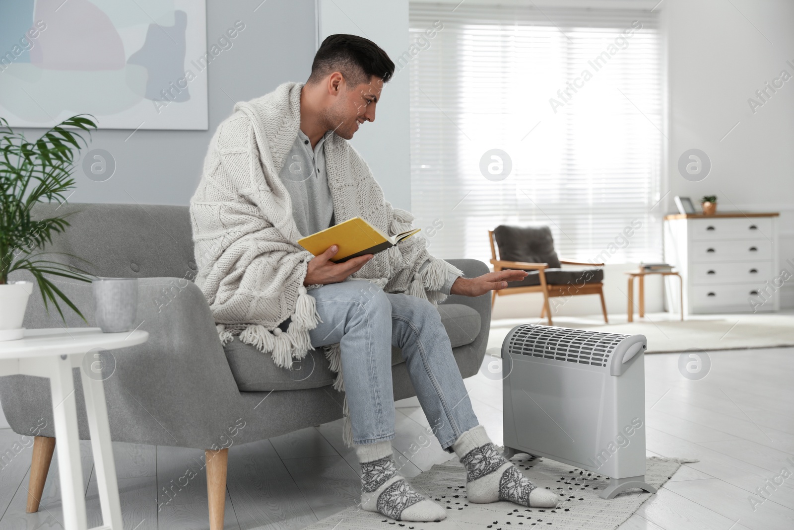 Photo of Happy man with book sitting on sofa near electric heater at home