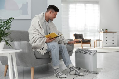 Photo of Happy man with book sitting on sofa near electric heater at home