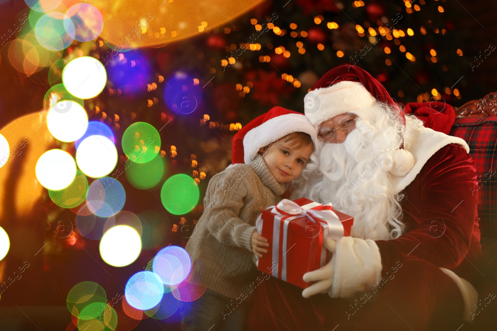 Photo of Santa Claus and little boy with gift near Christmas tree indoors