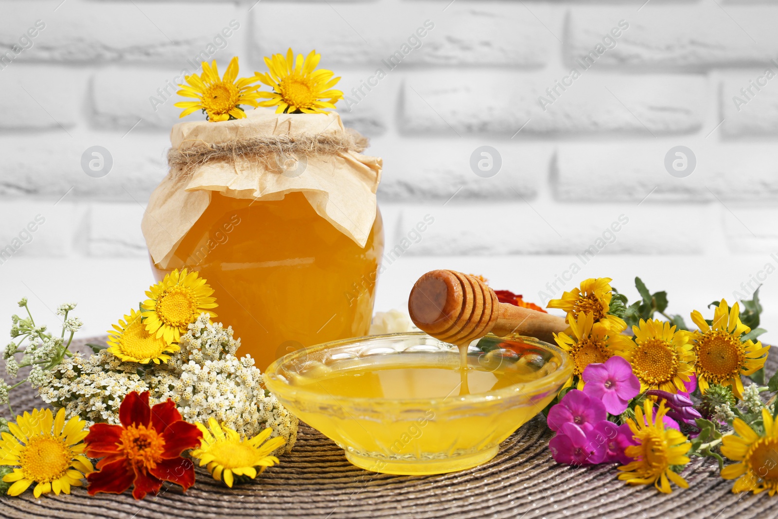 Photo of Delicious honey and dipper with different flowers on wicker mat near white brick wall