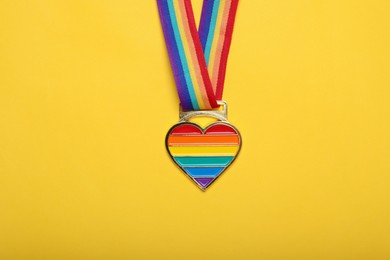 Photo of Rainbow ribbon with heart pendant on yellow background, top view. LGBT pride