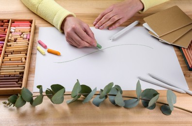 Photo of Woman drawing eucalyptus branch with soft pastels at wooden table, closeup