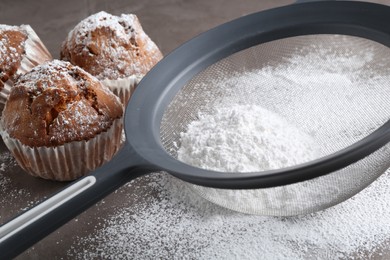 Photo of Sieve with sugar powder and muffins on grey table, closeup