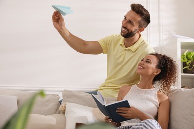 Photo of Happy man playing with paper plane while his girlfriend reading book on sofa in living room