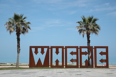 Photo of BATUMI, GEORGIA - JUNE 10, 2022: Beautiful art installation with word WHERE and palm trees near sea on sunny day