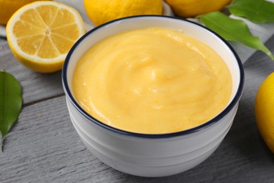 Delicious lemon curd in bowl, fresh citrus fruits and green leaves on grey wooden table, closeup
