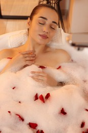 Photo of Woman taking bath in tub with foam and rose petals indoors