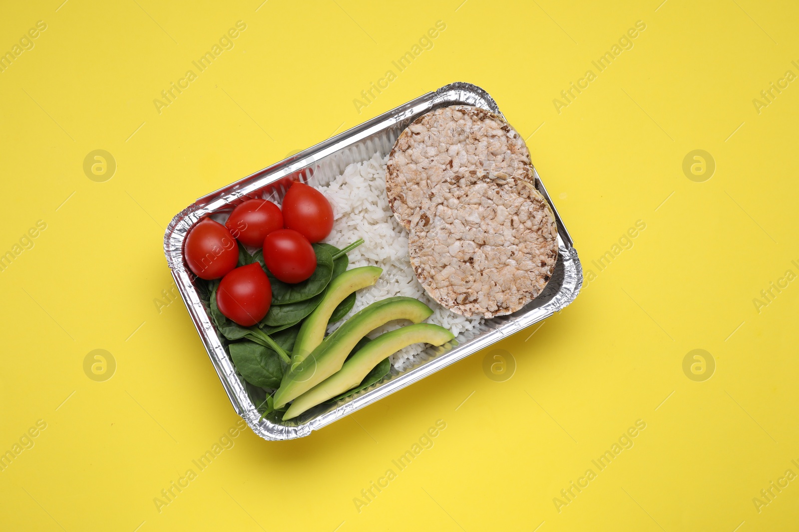 Photo of Container with rice, puffed crunchy cakes, fresh tomato, avocado and spinach on yellow background, top view