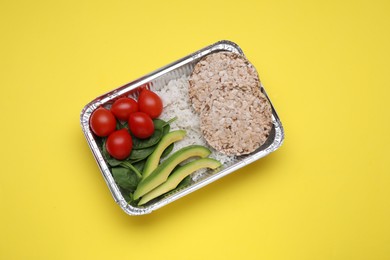 Container with rice, puffed crunchy cakes, fresh tomato, avocado and spinach on yellow background, top view