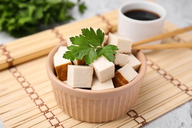 Photo of Bowl of smoked tofu cubes, soy sauce and parsley on bamboo mat, closeup