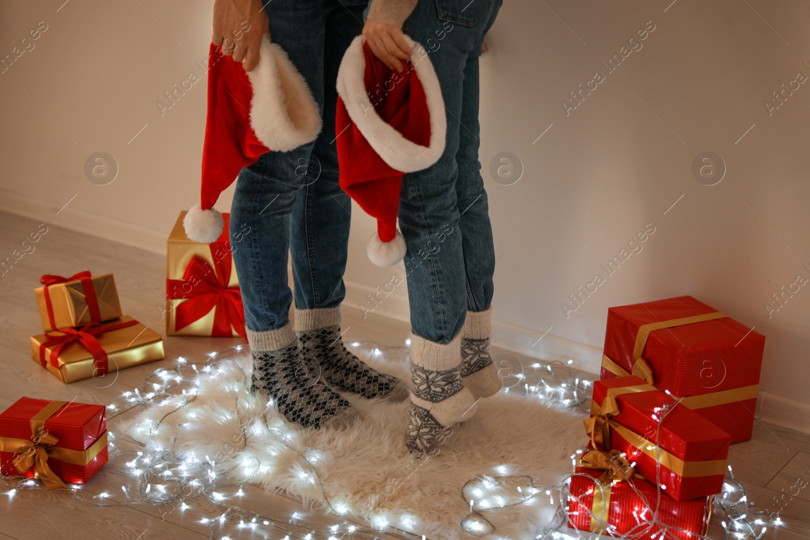 Photo of Young couple with Santa hats standing on rug near Christmas lights and gift boxes, closeup