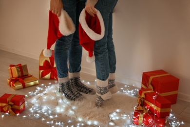 Photo of Young couple with Santa hats standing on rug near Christmas lights and gift boxes, closeup