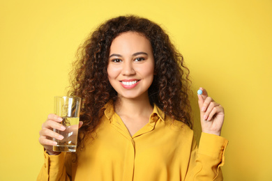 Photo of African-American woman with glass of water and vitamin pill on yellow background