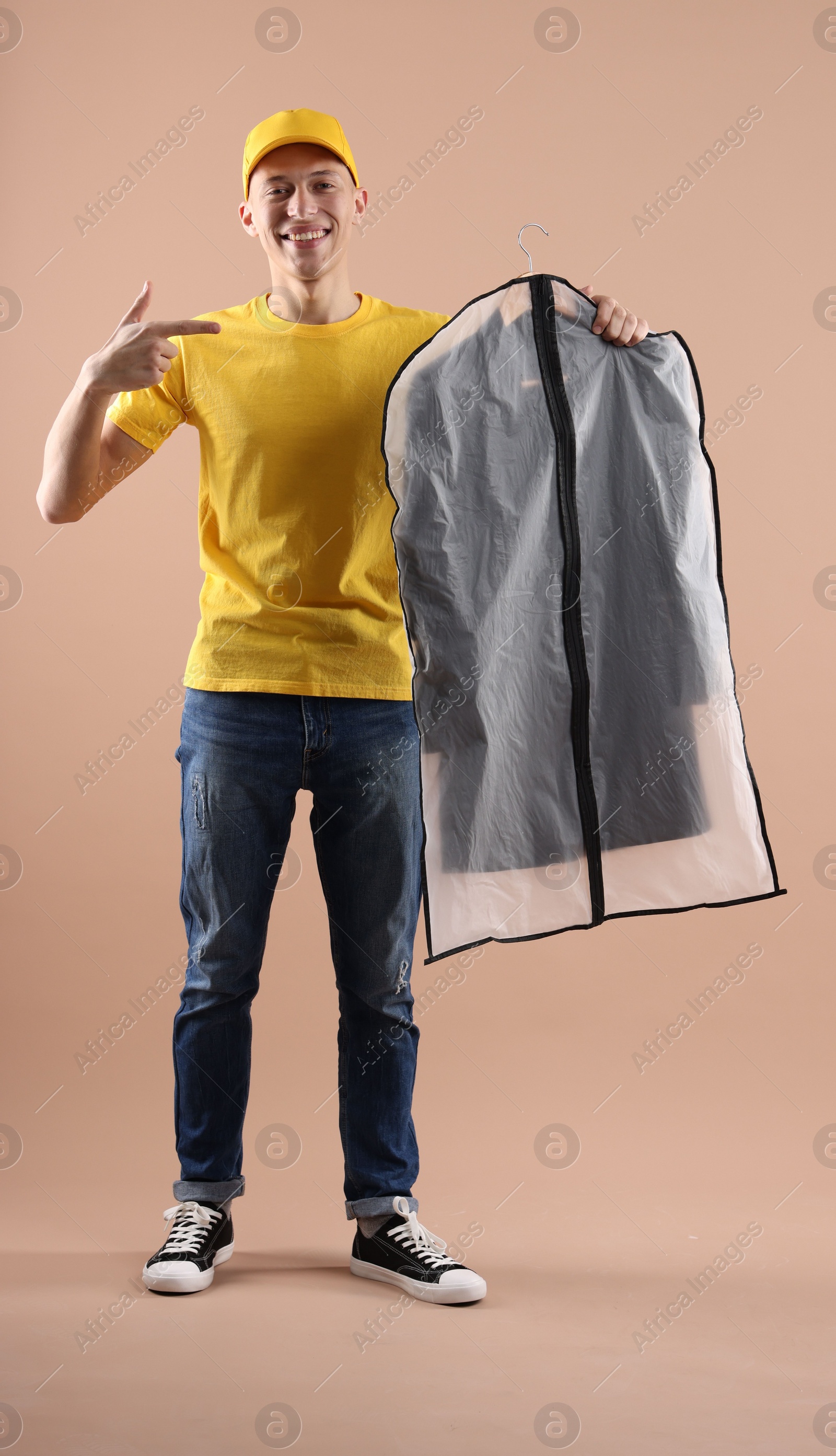 Photo of Dry-cleaning delivery. Happy courier holding garment cover with clothes on beige background